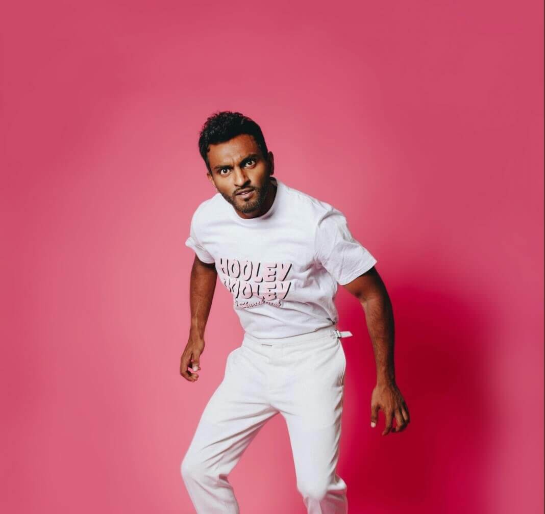 Comedian Nazeem Hussain jumping into the air in front of a pink background.