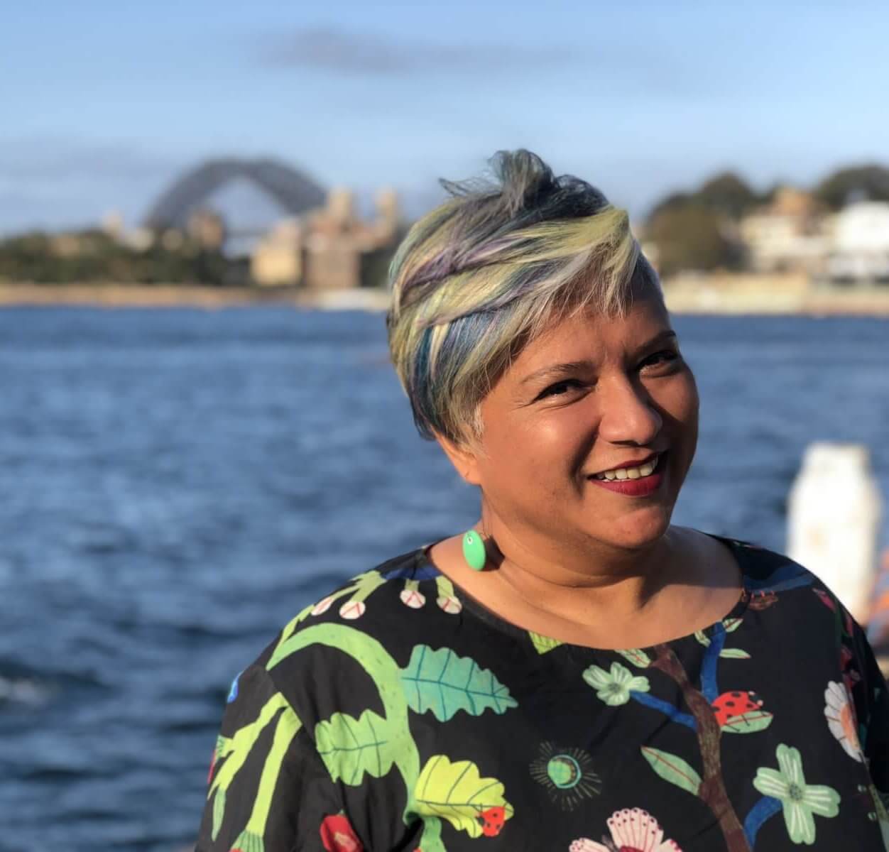 An image of Dr. Ruth De Souza near the sea, smiling at the camera. She is wearing a black blouse with multi coloured patterns on it