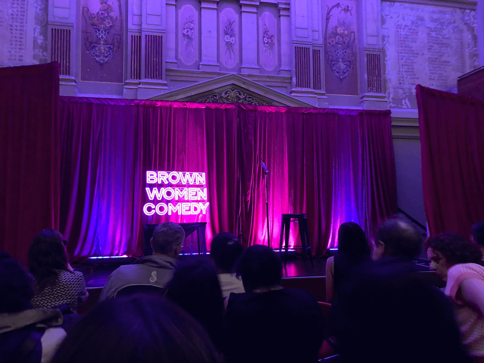 A photo of the Brown Women Comedy set. A bright pink neon sign saying 'BROWN WOMEN COMEDY' sits next to a microphone in front of red stage curtains on a stage set up in the middle of an old building with classical art on its walls.